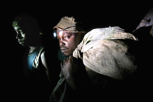 金鉱石が詰まった約３０キロの袋を軽々と担ぎながら、坑道の暗闇に浮かび上がる地下鉱山の労働者ら＝コンゴ民主共和国東部州モングワルのマカラ金鉱山で２００９年６月２８日、森田剛史撮影