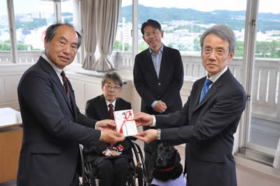 宝塚市ゴルフ場協議会の小堂剛裕会長（左）から寄付金の目録を受け取る毎日新聞大阪社会事業団の森野茂生常務理事（右）。（後方左から）木村佳友さんと森臨太郎市長＝同市役所で２０２５年５月２７日午後５時２５分、関谷徳撮影